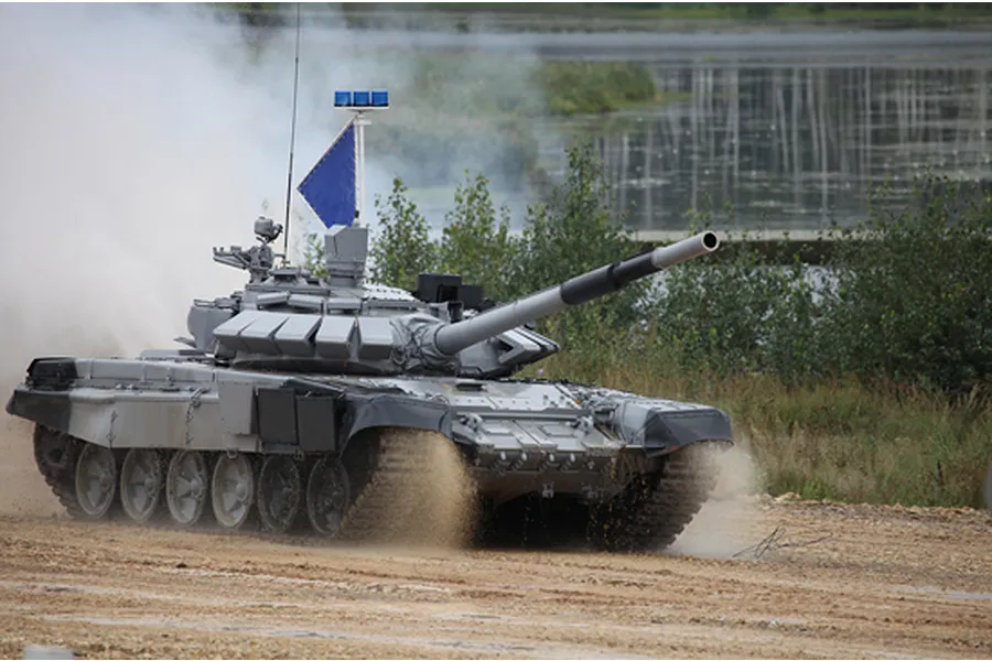 A Russian T-72B3M main battle tank driving through a dusty field with a blue flag and lights mounted on the turret.