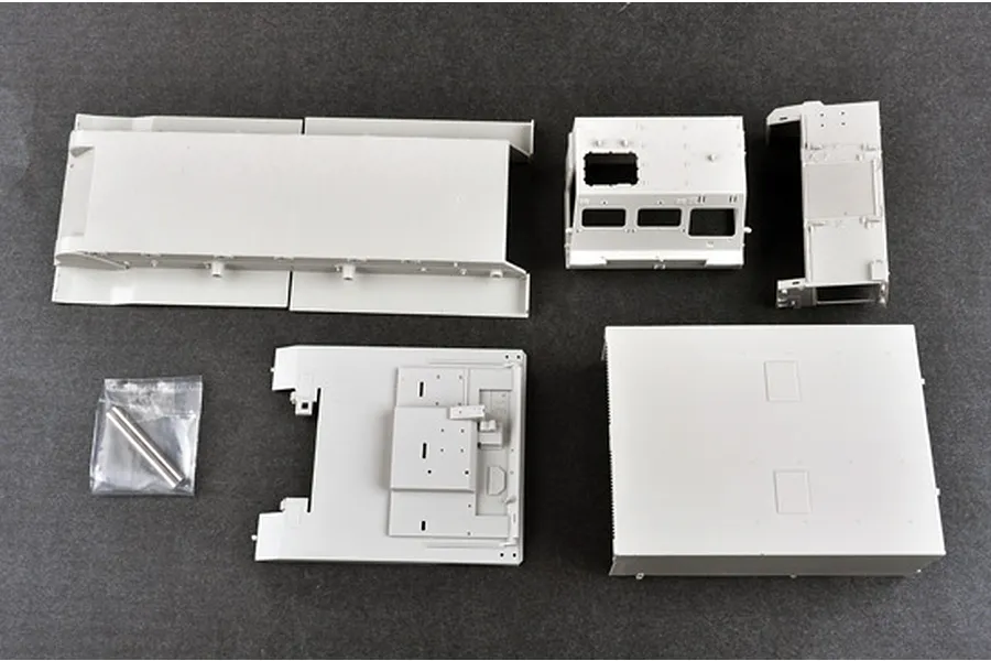 Unassembled grey plastic model kit parts for a military vehicle, including chassis and cabin components, laid out on a dark grey surface.