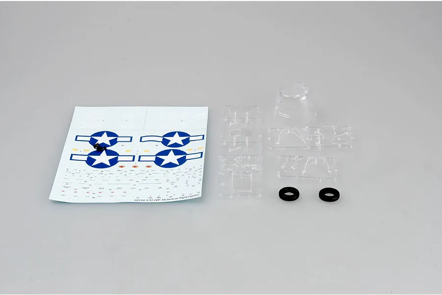 A decal sheet and clear plastic model kit parts including a canopy and two small black rubber rings on a plain grey background.