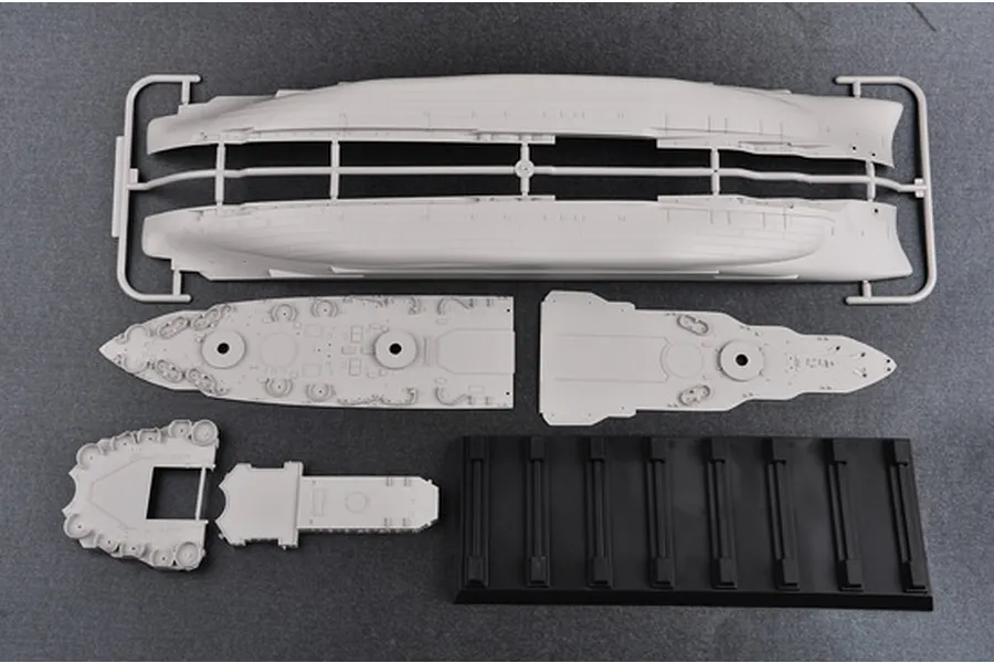 Plastic model kit parts for a battleship, including hull sections, deck pieces, and a black display stand on a grey surface.