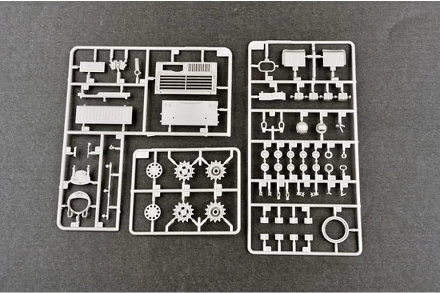 Three grey plastic model kit sprues containing various tank parts, including wheels, sprockets, and hull panels, laid out on a dark surface.