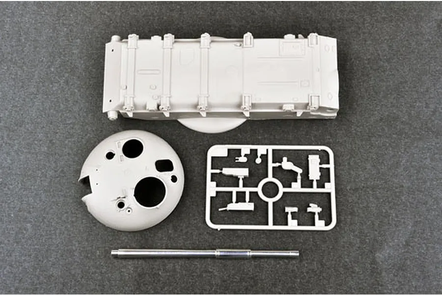 Top-down view of unpainted plastic model tank parts including the hull, turret, a small parts sprue, and a metal gun barrel on a dark grey surface.