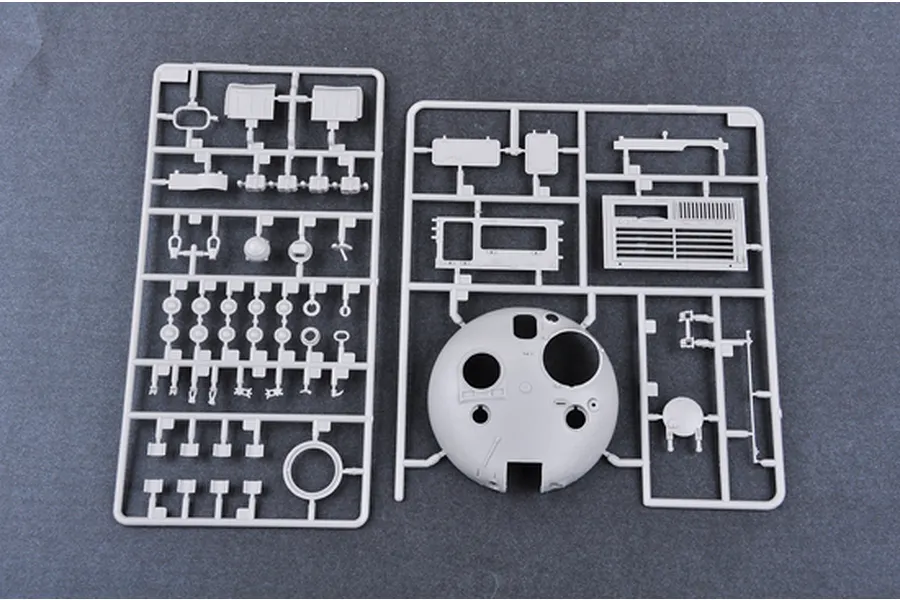 Two grey plastic model kit sprues containing various tank parts, including a large turret shell, laid out on a dark grey surface.