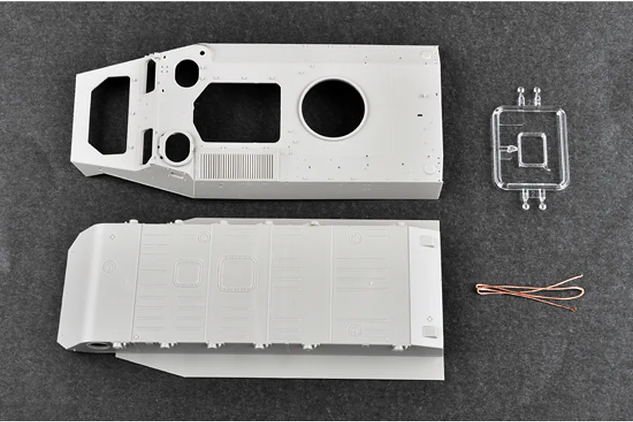 Top-down view of unpainted plastic model kit parts for a 1/35 scale military vehicle, including hull sections, a clear sprue, and a copper wire.
