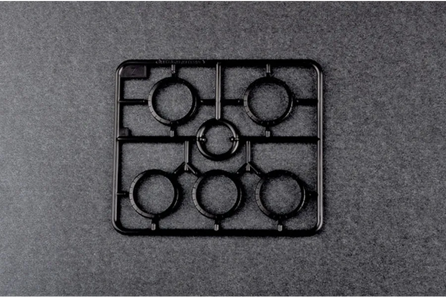 A black plastic sprue frame containing five circular ring parts for a model kit, viewed from above against a dark grey textured background.