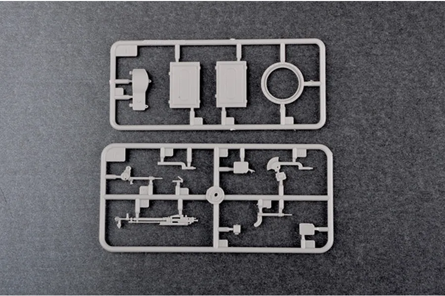 Two grey plastic model kit sprues containing various small tank parts, including a machine gun and hatch components, on a dark grey background.