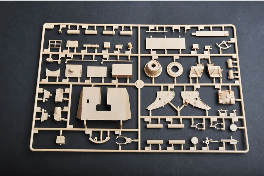 A plastic model kit sprue containing various tan-colored parts for a military vehicle model, laid out on a dark grey surface.