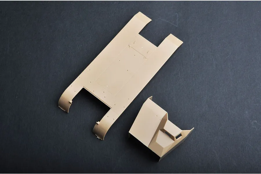 Tan plastic model kit parts for a military vehicle, including a chassis floor and cabin section, on a dark grey surface.