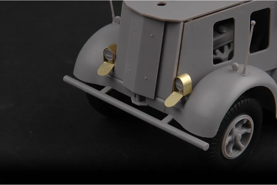Close-up of the front end of an unpainted grey plastic model half-track vehicle with brass headlight covers.
