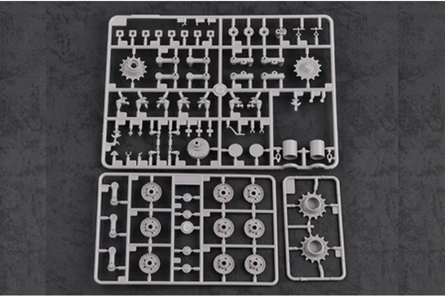 Three grey plastic model kit sprues containing various tank parts like wheels and sprockets on a dark textured background.