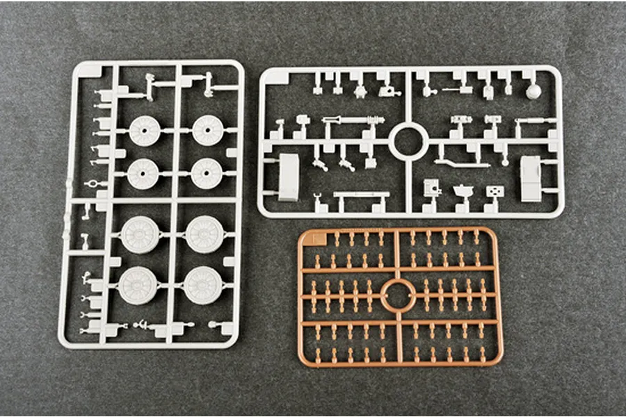 Three plastic model kit sprues on a dark grey background, including two light grey sprues with wheels and parts, and one tan sprue with small components.