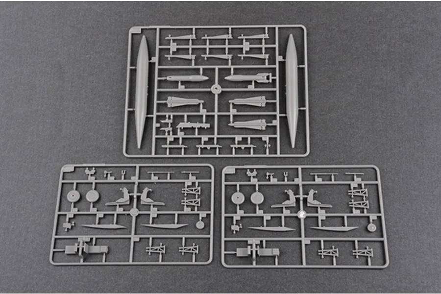 Three grey plastic model kit sprues containing aircraft parts like fuel tanks, seats, and landing gear components on a dark grey background.
