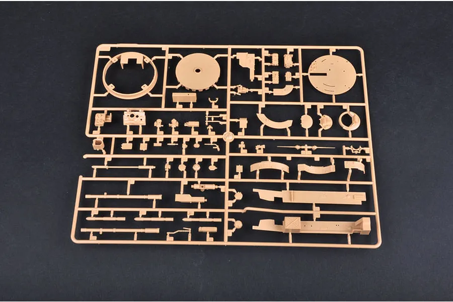 A tan plastic model kit sprue containing various tank parts, including turret components and gun barrels, against a dark grey background.