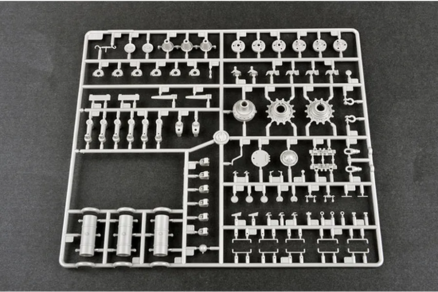 A grey plastic sprue containing various model tank parts, including wheels, fuel drums, and suspension components, laid out on a dark grey surface.