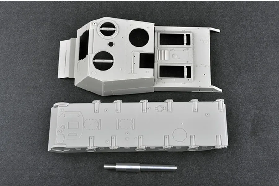 Unassembled grey plastic model tank hull, upper superstructure, and metal gun barrel on a dark grey surface.