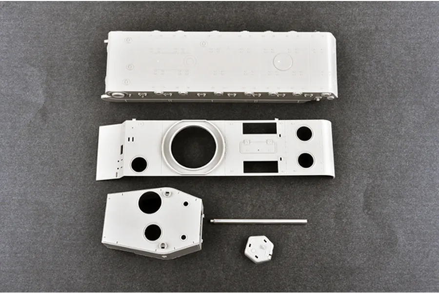 Unassembled grey plastic model kit parts for a tank, including the hull, turret, and gun barrel, laid out on a dark grey surface.
