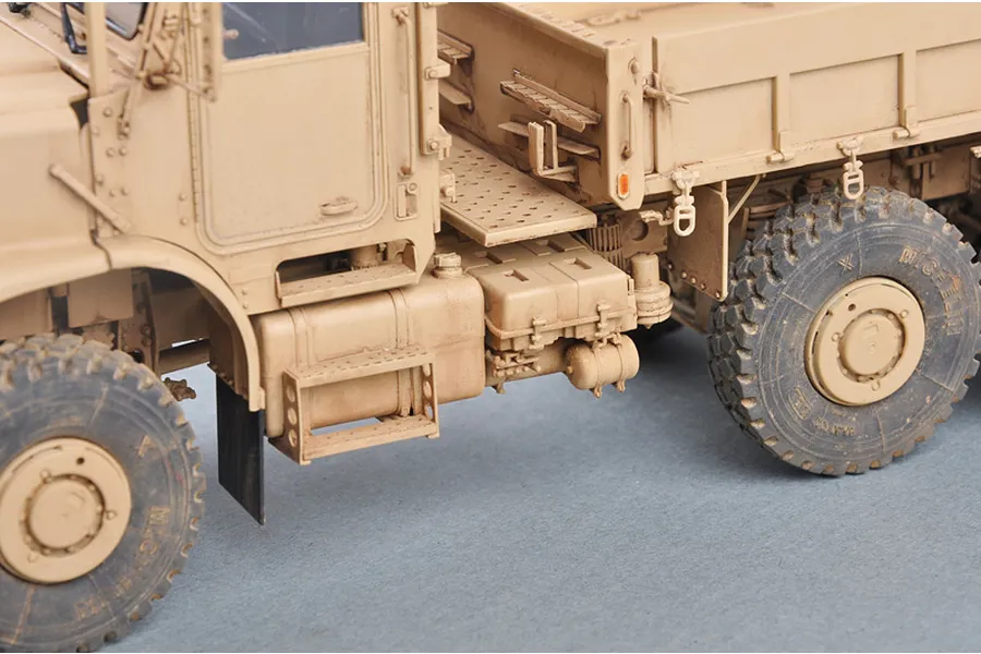A close-up side view of a 1/35 scale model of a tan U.S. MK.23 MTVR military truck, showing the cab, steps, fuel tank, and large off-road tires.