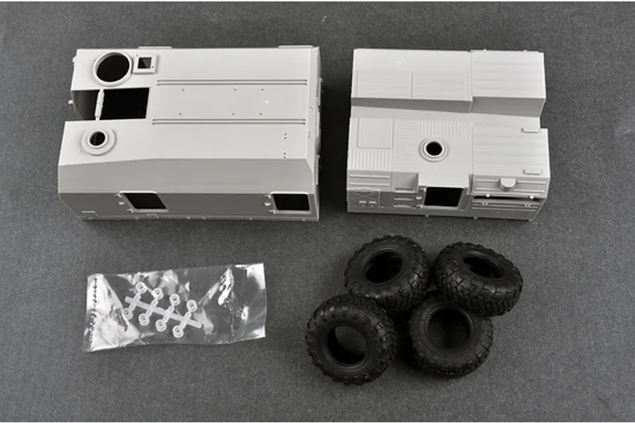 Unassembled plastic model kit parts for a military vehicle, including two large grey hull sections, four black rubber tires, and a small bag of clear plastic components.