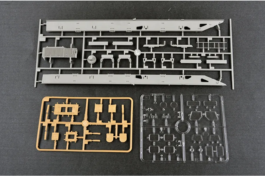 Three plastic model kit sprues on a dark grey background, including grey, tan, and clear plastic parts for a military vehicle model.