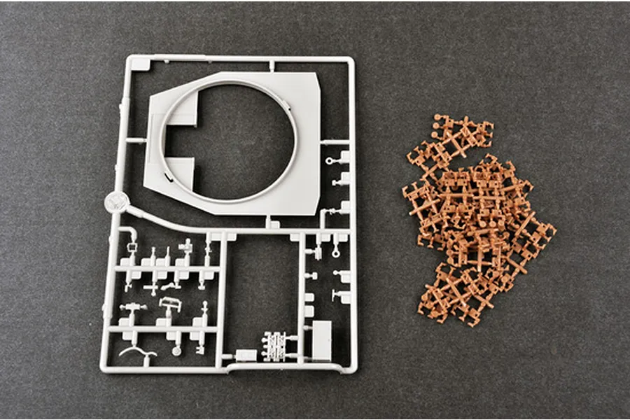 A grey plastic model kit sprue featuring a large turret ring and various small parts, alongside a pile of loose brown plastic tank track links.