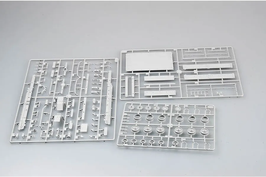 Three grey plastic model kit sprues containing various vehicle parts, including wheels and chassis components, laid out on a plain light grey background.