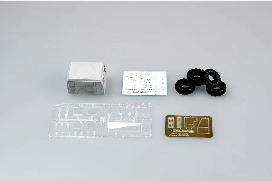 A selection of model kit parts for a Trumpeter 1/35 M1078 truck, including a cab section, decal sheet, rubber tires, clear plastic sprue, and photo-etched parts.