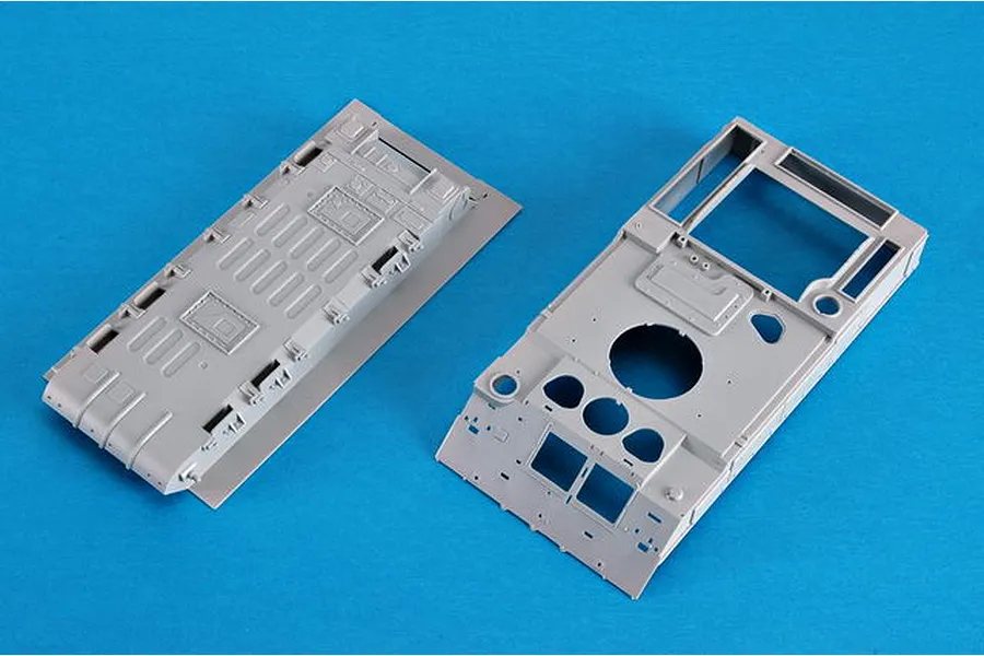 Two grey plastic model kit parts for a military vehicle, showing the upper and lower hull sections on a blue background.
