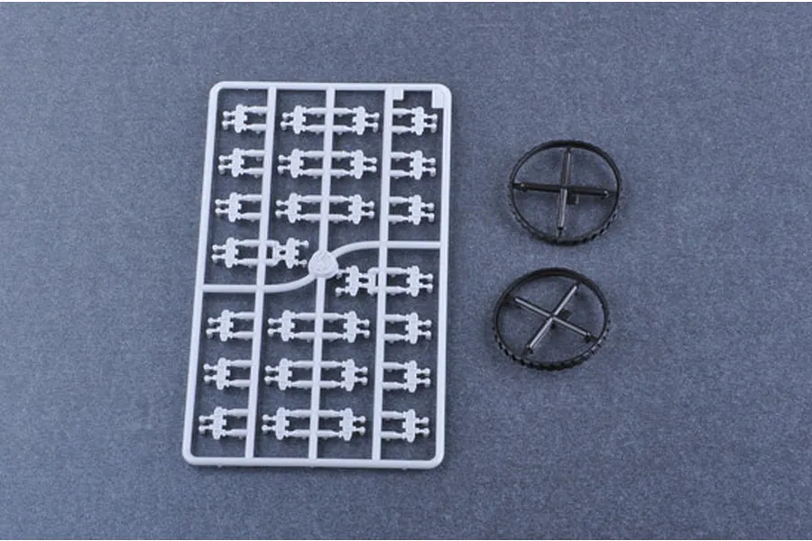 A plastic model kit sprue with small parts and two circular black wheels on a grey background.