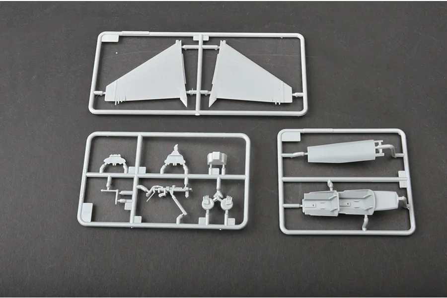 Three grey plastic model kit sprues containing aircraft parts like wings and cockpit components on a dark surface.