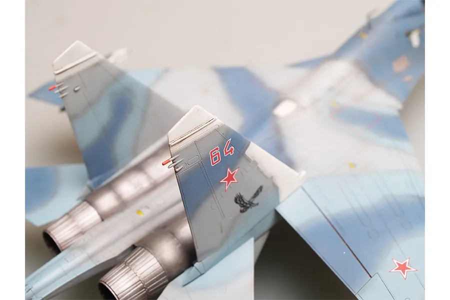 A close-up, high-angle view of a scale model Su-27 fighter jet, showing the tail fins with red number 64 and a red star emblem.