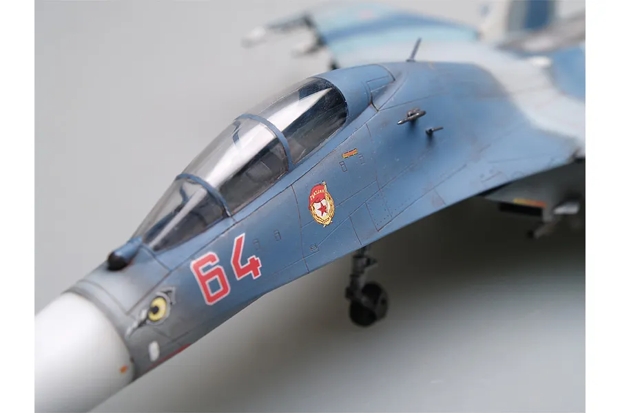 A close-up view of a scale model Su-27UB fighter jet, showing the cockpit, nose cone, and Soviet-style markings on a blue and grey camouflage fuselage.