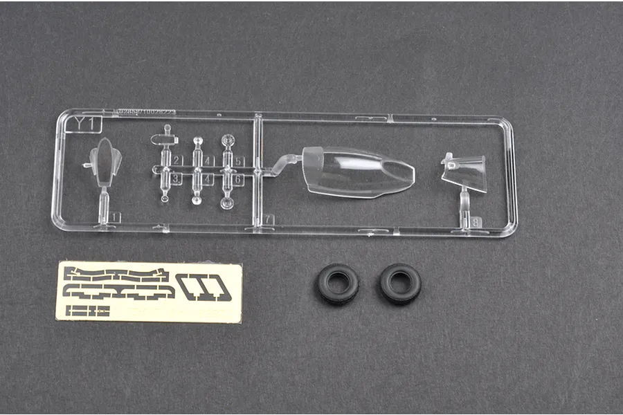 A clear plastic sprue containing canopy and small parts, a small sheet of masking tape, and two black rubber tires for a 1/48 scale model kit.