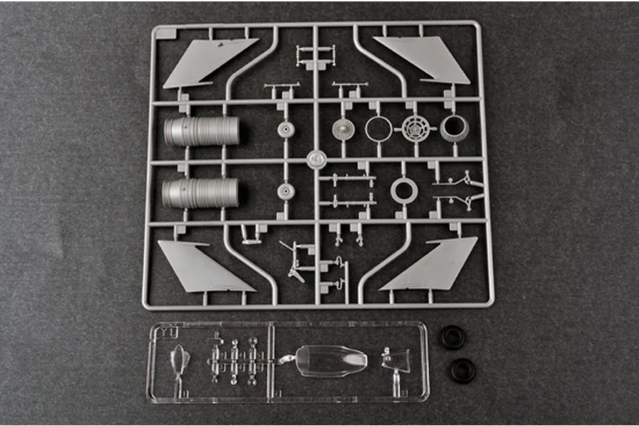 A grey plastic sprue containing aircraft model parts, a clear plastic sprue with canopy parts, and two black rubber tires on a dark grey surface.