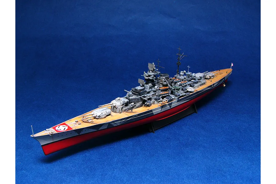 A fully assembled and painted 1/700 scale model of the German battleship Tirpitz, featuring a camouflage hull and wooden deck, displayed on a stand.