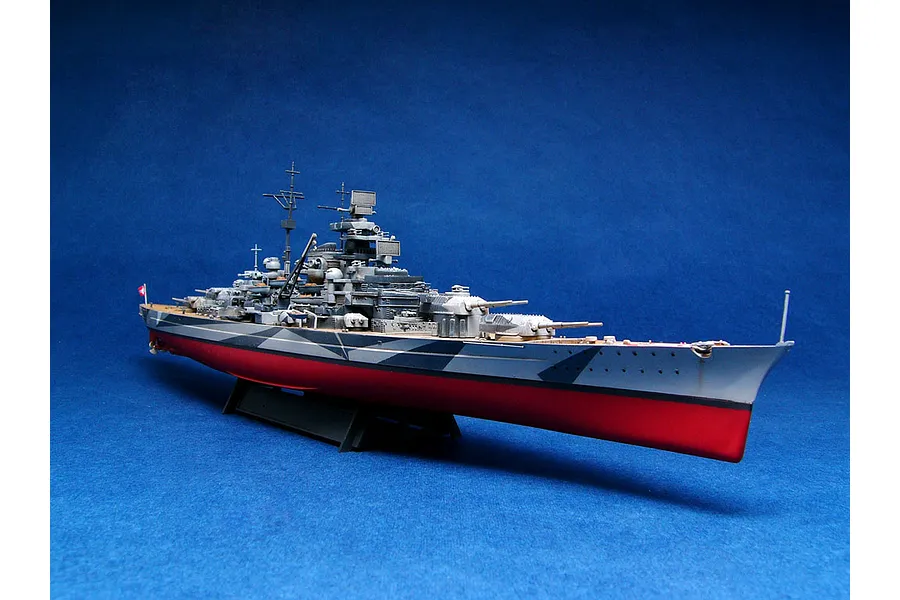 A fully assembled and painted 1/700 scale model of the German battleship Tirpitz, featuring a camouflage hull and red lower hull, displayed on a stand.