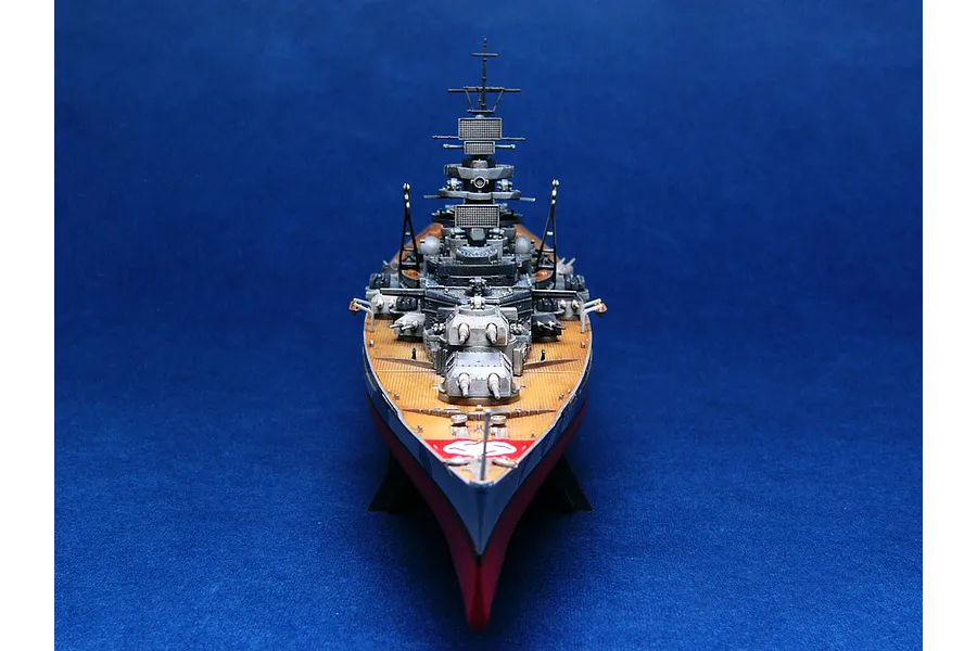 A front-facing view of a 1/700 scale model of the German battleship Tirpitz, featuring detailed deck, turrets, and superstructure on a blue background.