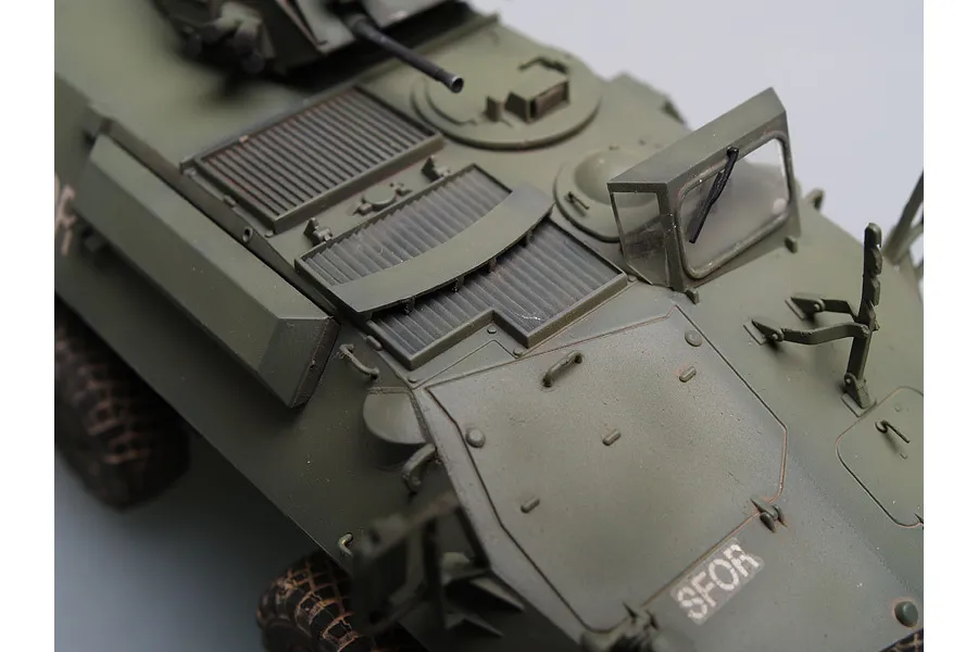A high-angle close-up of a 1/35 scale model of a Canadian Grizzly AVGP armoured vehicle, showing the olive green hull, engine deck, and SFOR markings.