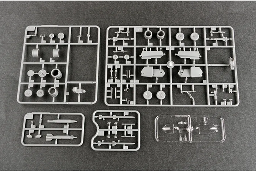 Four grey plastic model kit sprues and one clear plastic sprue containing aircraft parts laid out on a dark surface.