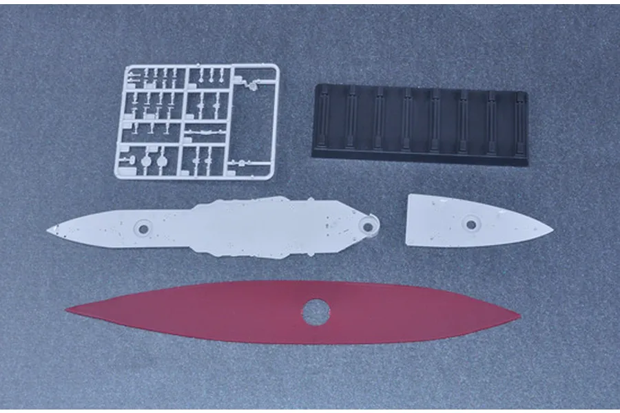 Top-down view of plastic model kit parts for a battleship, including a grey sprue, a black display stand, and grey and red hull sections.