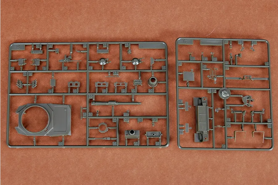 Two grey plastic model kit sprues containing various tank parts, including a turret top and main gun barrel, laid out on a brown surface.