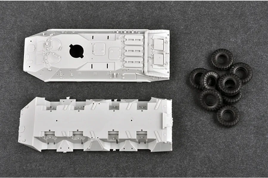 Unassembled plastic model kit parts for a BTR-70 APC, including the upper hull, lower hull, and several black rubber tires on a dark grey surface.