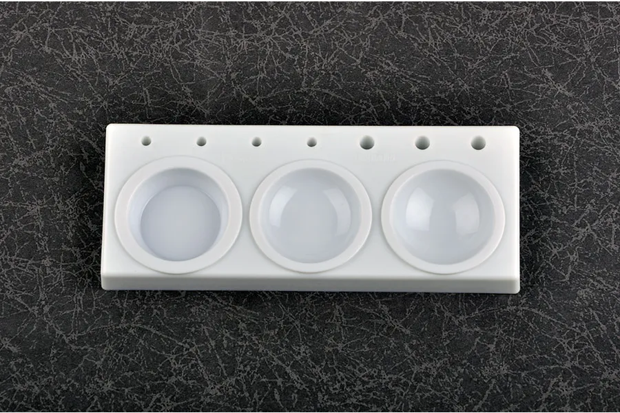 A white plastic Trumpeter modeler's paint palette with three circular mixing wells and seven small holes for holding brushes, set against a dark textured background.