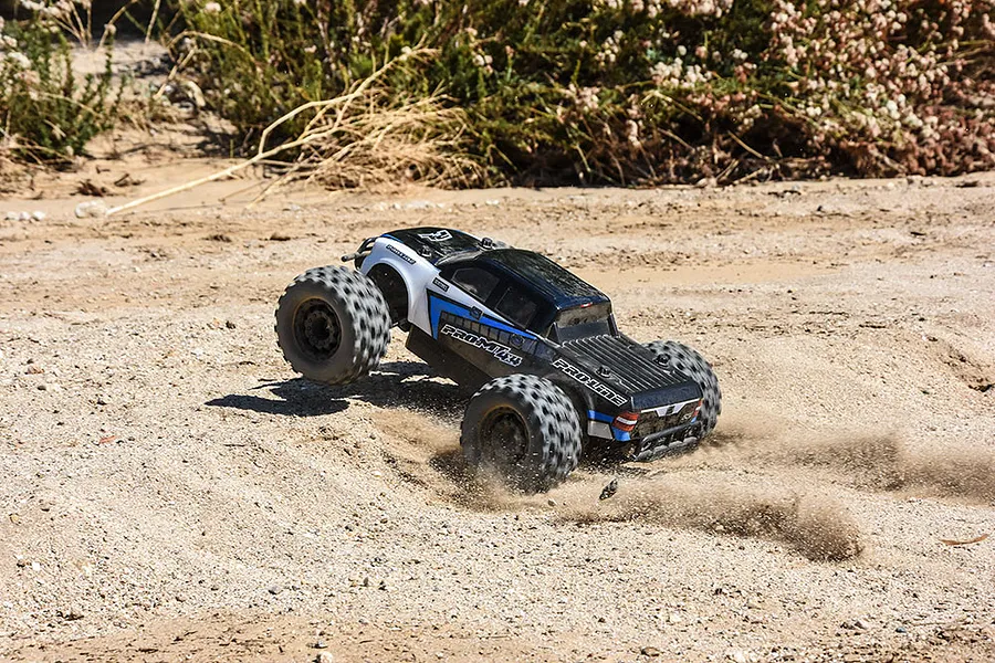 A Pro-Line PRO-MT 4x4 RC monster truck driving over sandy, uneven terrain, kicking up dust.