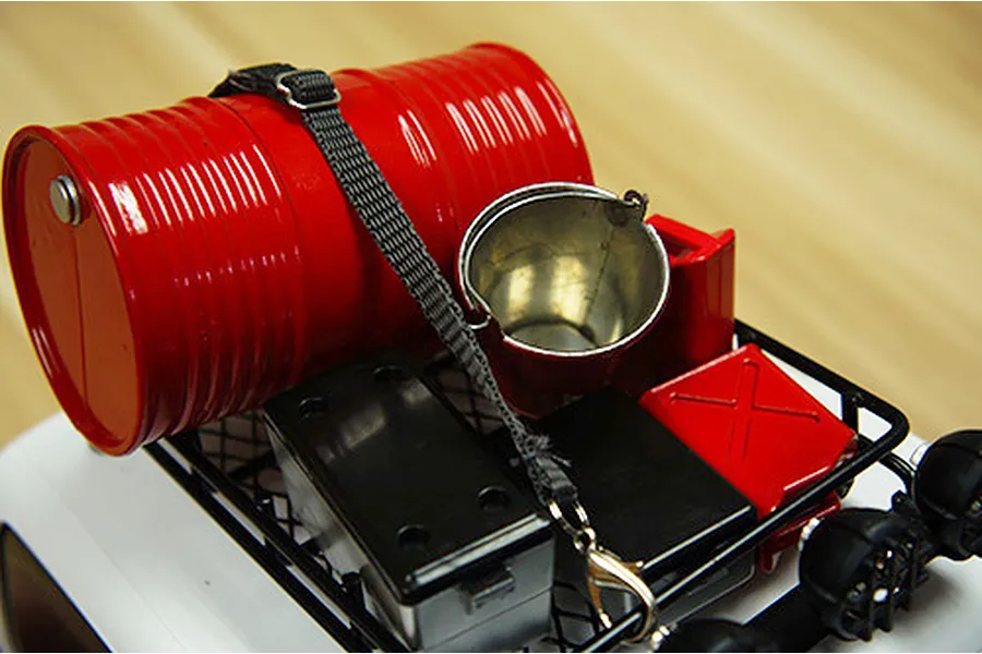 A black nylon cable strap with a metal buckle and spring-loaded hook securing a red scale fuel drum to an RC car roof rack.