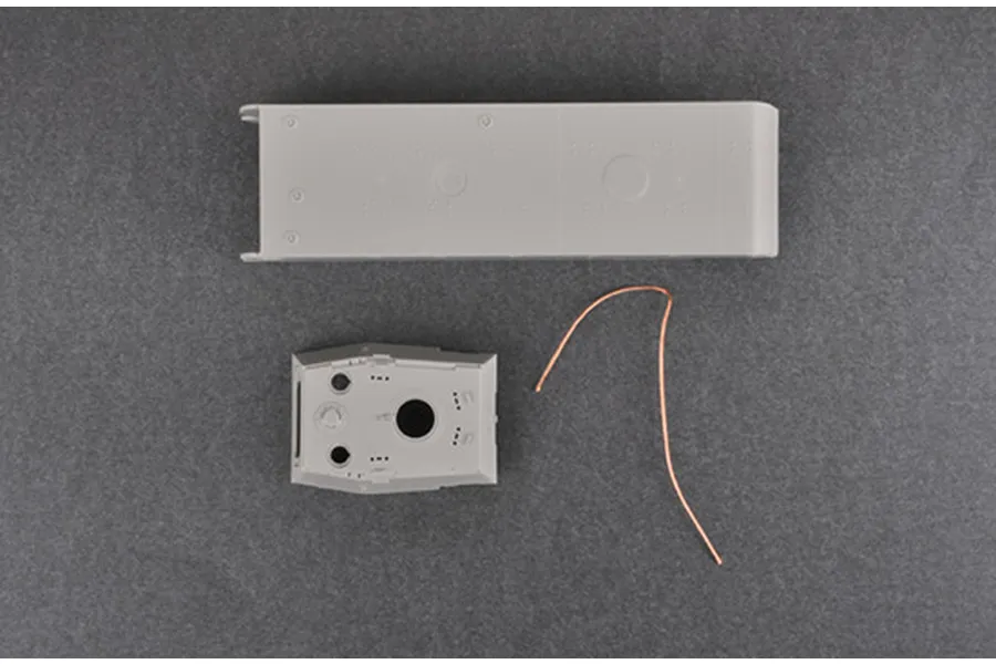 Plastic model tank parts including a hull section, turret top, and a piece of copper wire on a grey background.