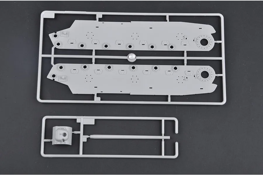 Two grey plastic model kit sprues containing tank hull sides and a gun barrel piece on a dark surface.