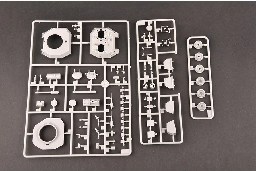 Three grey plastic model kit sprues containing various vehicle parts, including turret components and wheels, laid out on a dark surface.