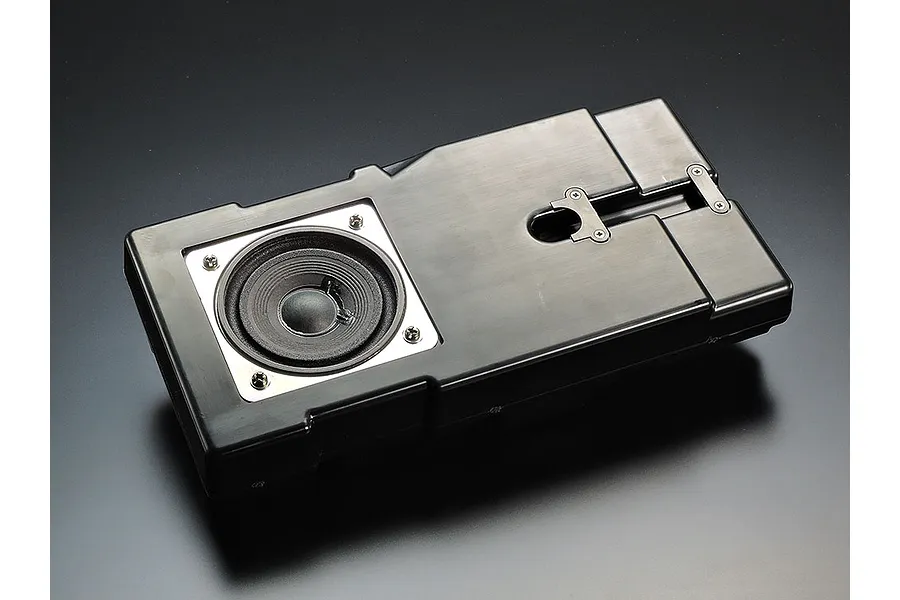 A black plastic speaker box component for a Tamiya RC tank kit, featuring a circular speaker driver mounted with metal screws.