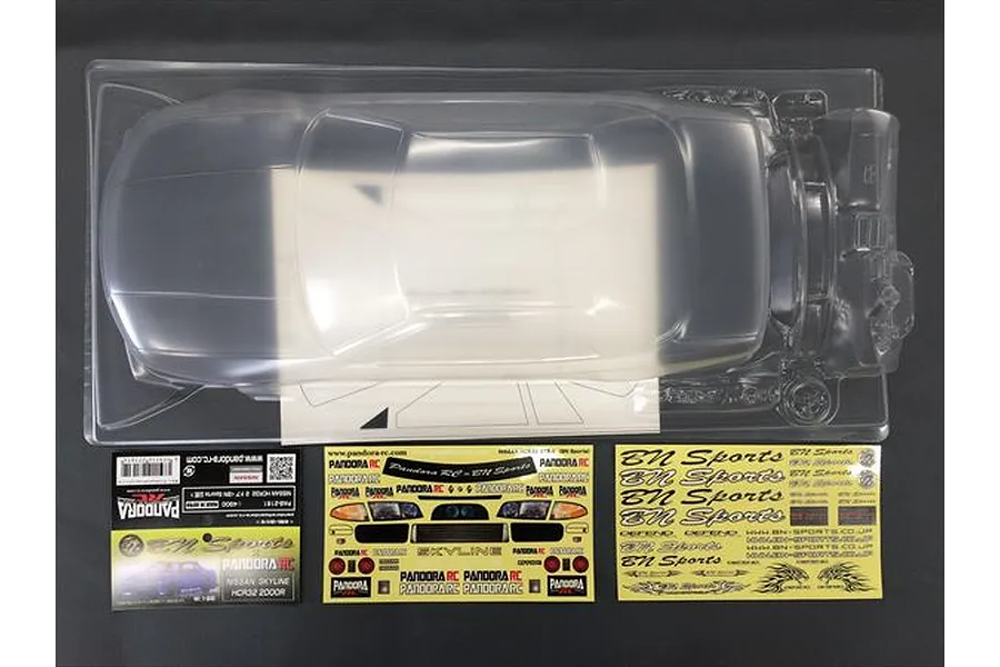 Unpainted clear polycarbonate RC car body shell for a Nissan HCR32, including decal sheets and window masks in a plastic tray.