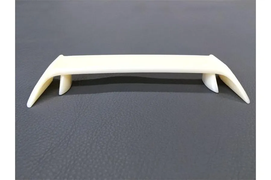 An unpainted white plastic RC car rear wing spoiler sitting on a dark grey textured surface.
